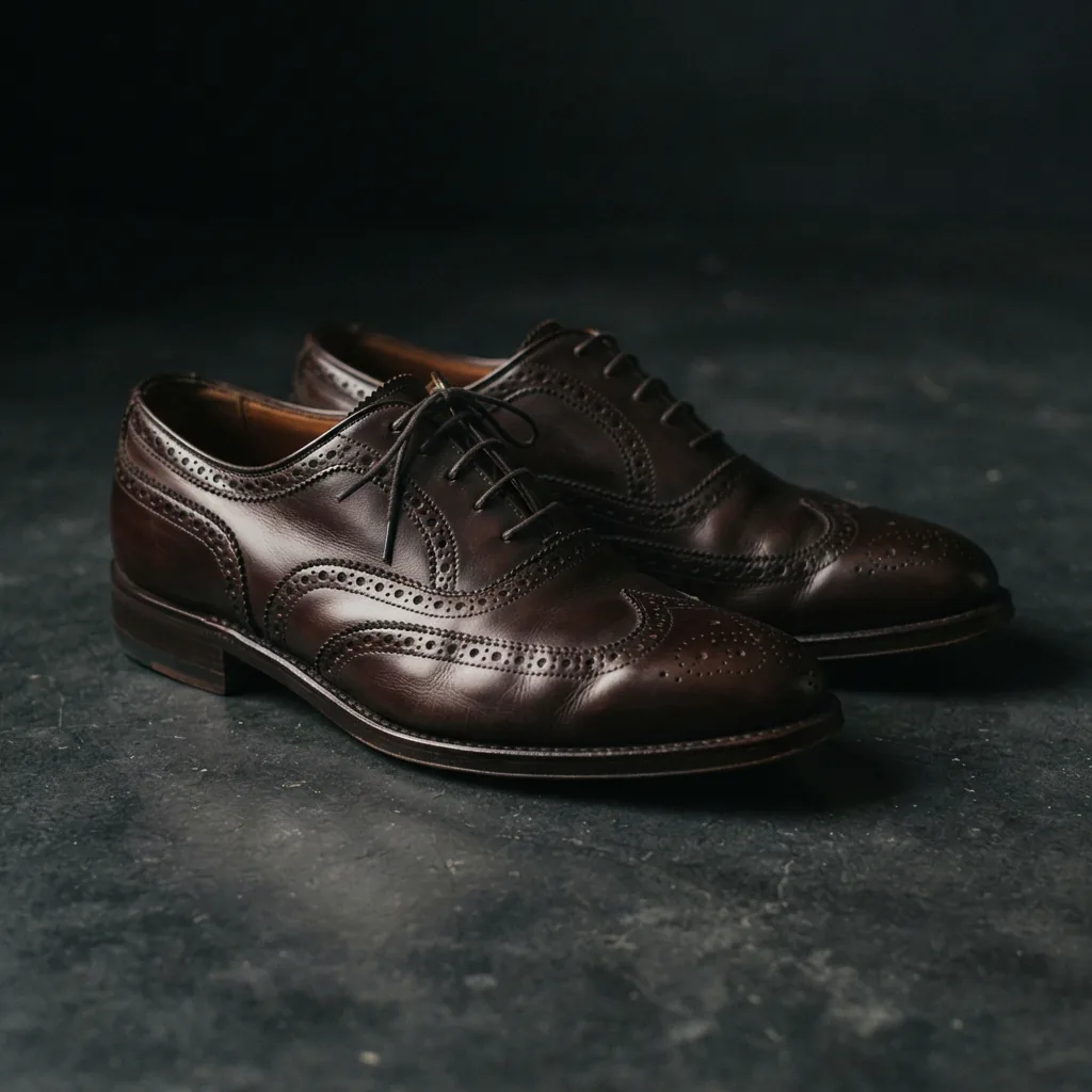 Two men's dress shoes with wingtip and brogue detailing on a dark studio surface.