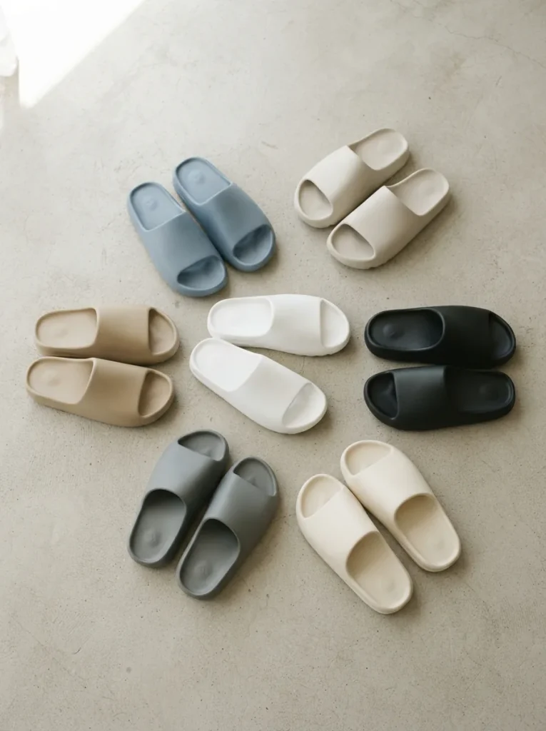 Overhead arrangement of assorted unbranded slide sandals in mixed neutral tones on pale concrete